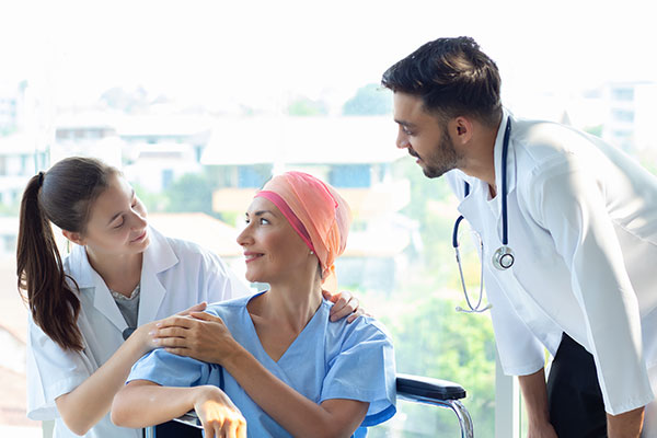 Doctor and nurse taking care of a senior Caucasian woman with a head cover caused by chemotherapy cancer, smiling and sitting in a wheelchair.
